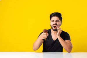 Indian young man eating ice cream in cone and having teeth ache because of cavity, sitting at table against yellow or white background